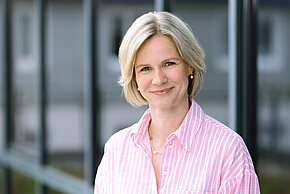 Frau mit kurzem blonden Harren und einer pinken bluse lächelt in die Kamera.