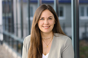 Frau mit langen braunen Haaren und hellem Oberteil lächelt in die Kamera.