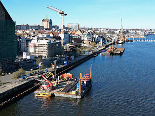 Blick auf einen Hafen mit Bauarbeiten auf Pontons im Wasser und Kränen, im Hintergrund Stadt mit Gebäuden und blauem Himmel