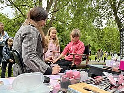 Frau malt einem sitzenden Mädchen im Park ein Tattoo, umgeben von weiteren Kindern und Erwachsenen