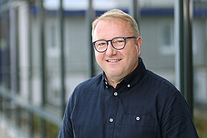 Mann mit Brille und dunklem Hemd sitzt vor einem Fenster.