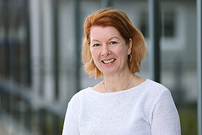 Frau mit kurzen rotbraunen Haaren und einem weißen Oberteil gekleidet, lächelt in die Kamera.