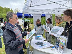 Mehrere Personen stehen unter einem weißen Pavillon und betrachten Fotografien, im Hintergrund grüne Wiese und Bäume