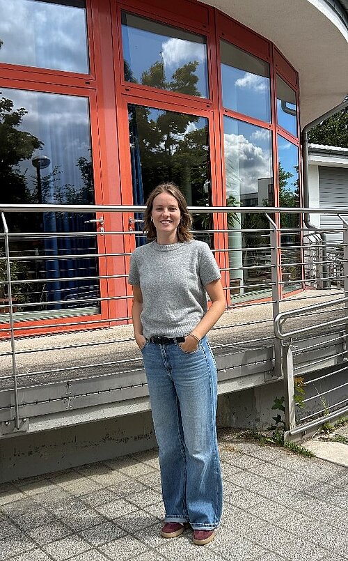 Frau im grauen T-Shirt und blauer Hose steht vor einem Fenster und lächelt in die Kamera.
