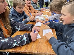 Kinder und Erwachsene sitzen an einem Holztisch und lösen gemeinsam Bingo auf Papier mit Stiften
