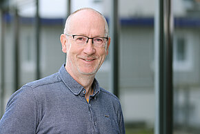 Mann mit Brille, trägt ein Hemd und lächelt in die Kamera.