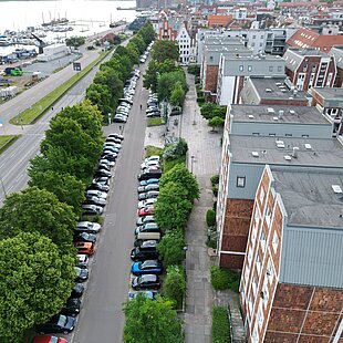 Blick von oben auf eine Straße mit parkenden Autos und Bäumen entlang eines Gewässers in einer Stadt.
