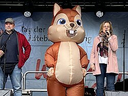 Zwei Personen stehen auf einer Bühne neben einer großen, braunen Eichhörnchen-Maskottchenfigur mit großen Augen und breitem Lächeln
