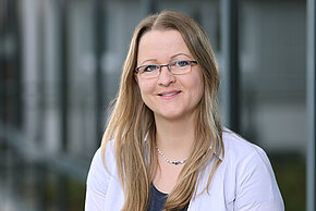 Frau mit langen Haaren und Brille, trägt einen blauen Blazer. Sie steht vor einer modernen Glaswand.