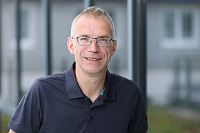 Mann mit Brille und kurzem, glattem Haar, trägt ein dunkles Poloshirt und lächelt in die Kamera.