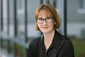 Eine Frau mit kurzen braunen Haaren und Brille, die in eine schwarze Bluse trägt und in die Kamera lächelt.