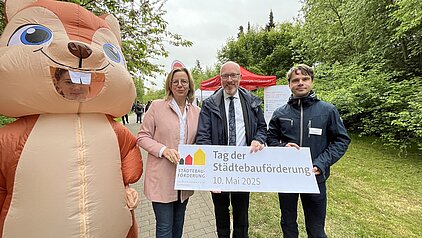 Drei Personen und eine Person im Eichhörnchen-Kostüm halten ein Schild mit der Aufschrift 'Tag der Städtebauförderung 10. Mai 2025' in einem Park mit Bäumen und einem roten Zelt im Hintergrund.