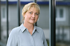 Porträt einer Frau mit blonden Haaren, die in einem blauen Oberteil gekleidet ist, vor einer Glaswand.