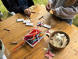 Kinder sitzen an einem Holztisch und bemalen ausgeschnittene Schmetterlingsformen aus Papier mit Buntstiften und Filzstiften.