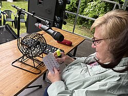 Ältere Frau mit Brille und grauer Jacke hält Bingo-Karte und Kugel, sitzt an Tisch mit Bingo-Maschine und Mikrofon