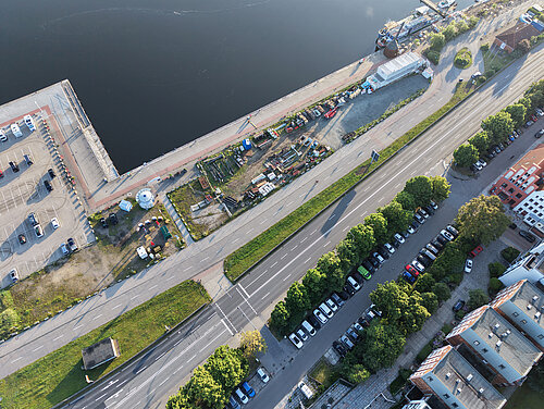 Luftaufnahme eines Flussufers mit angrenzender Straße, Parkplätzen, Bäumen und Gebäuden