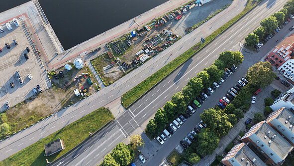 Luftaufnahme eines Flussufers mit angrenzender Straße, Parkplätzen, Bäumen und Gebäuden