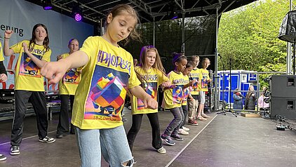 Kinder tanzen auf einer Bühne bei einer Veranstaltung, tragen gelbe T-Shirts mit buntem Aufdruck und bewegen synchron ihre Arme.
