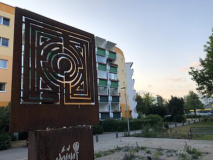 Metalltafel mit Labyrinthmuster vor Wohngebäuden und Bäumen im Freien.