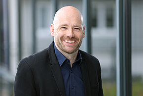 Mann mit Glatze und Bart, trägt ein schwarzes Sakko und ein blaues Hemd und lächelt.