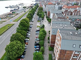 Strandstraße (Westabschnitt) Blick von oben auf eine Straße mit parkenden Autos und Bäumen entlang eines Gewässers in einer Stadt.