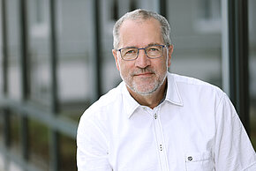 Mann mit Brille und grauem Bart, trägt ein weißes Hemd und lächelt.