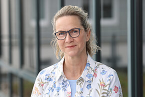Frau mit blonden Haaren und Brille, trägt eine weiße Bluse mit Blumenmuster und lächelt.