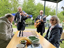 Zwei Musiker in Anzügen spielen Geige und Gitarre im Freien, während zwei ältere Personen an einem Tisch Schach spielen