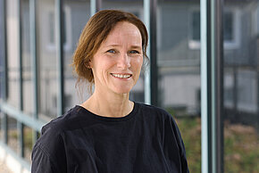 Frau mit braunen Haaren und dunklem Oberteil lächelt in die Kamera.