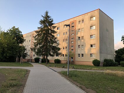 Fünfgeschossiges Wohngebäude mit beigen Fassaden und mehreren Fenstern, umgeben von Bäumen und einem Gehweg im Vordergrund.