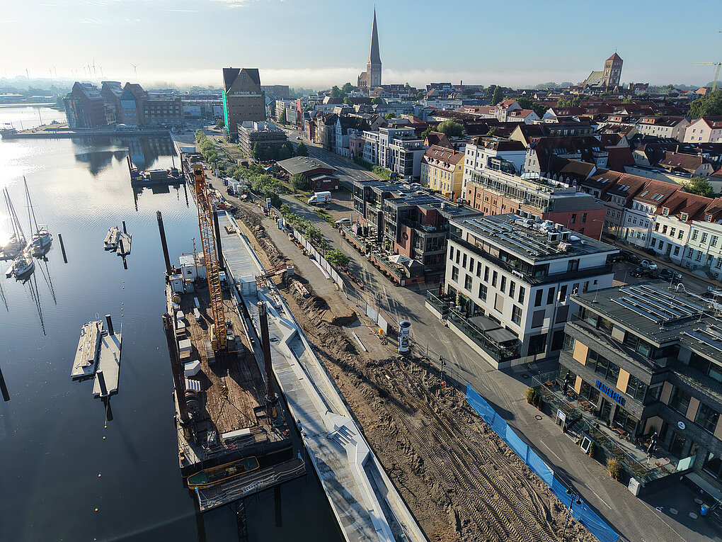 Luftaufnahme eines Stadthafens mit Baustelle entlang des Ufers und angrenzender Stadt mit Kirchturm im Hintergrund