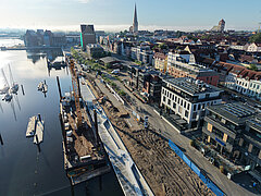 Luftaufnahme eines Stadthafens mit Baustelle entlang des Ufers und angrenzender Stadt mit Kirchturm im Hintergrund