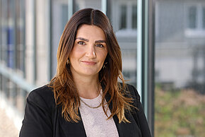 Frau mit langen braunen Haaren, hellen Oberteil und schwarzen Blazer lächelt in die Kamera.