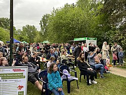 Große Gruppe von Menschen unterschiedlichen Alters sitzt und steht bei einer Outdoor-Veranstaltung mit Informationsständen und grünen Bäumen im Hintergrund