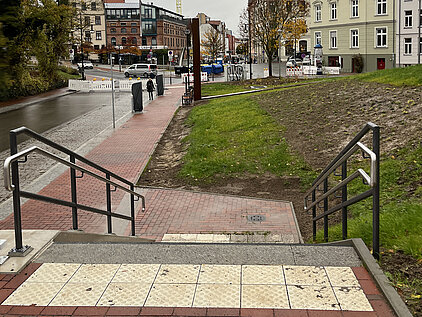 Blick von einer Treppe mit taktilen Bodenplatten auf eine Straße mit Baustellenabsperrungen, Bäumen und mehrstöckigen Wohnhäusern im Hintergrund