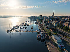 Luftaufnahme eines Stadthafens mit Segelbooten, Uferpromenade und Stadt mit Kirchturm im Hintergrund