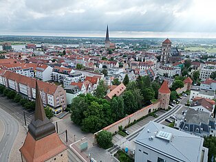 Fläche am Steintor Luftaufnahme der Rostocker Innenstadt mit roten Dächern, zwei markanten Kirchtürmen und einem Fluss im Hintergrund unter bewölktem Himmel