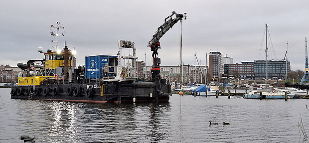 Arbeitsboot mit Kran und Containern im Stadthafen, im Hintergrund Segelboote und Gebäude