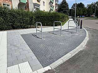 Drei silberne Fahrradständer auf gepflastertem Boden vor einem Wohnhaus mit Hecke