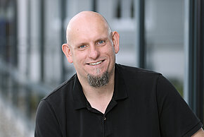 Mann mit Glatze und Bart, trägt ein schwarzes T-shirt und lächelt.