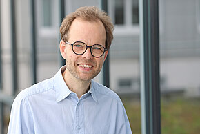 Mann mit Brille und hellem Hemd lächelt vor einer Fensterfront.