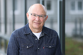 Mannmit Brille und kurzen, grauen Haaren, trägt ein dunkelblaues Jeanshemd und lächelt in die Kamera.