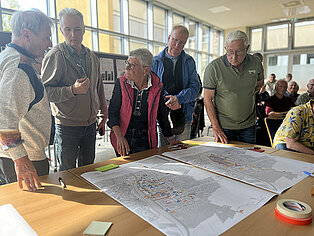 Mehrere Personen stehen um einen Tisch mit großen Plänen und diskutieren in einem hellen Raum mit großen Fenstern