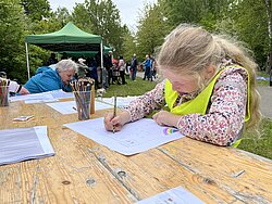 Mädchen mit gelber Weste sitzt an Holztisch im Freien und malt auf Papier, im Hintergrund Menschen und grünes Zelt