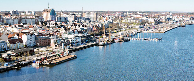 Blick auf einen Stadthafen mit mehreren Booten und Kranen, umgeben von Gebäuden und der Innenstadt im Hintergrund bei klarem Himmel