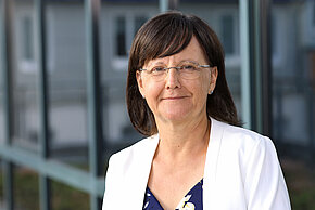 Frau mit kurzen braunen Haaren und Brille, trägt ein helles Oberteil und lächelt.