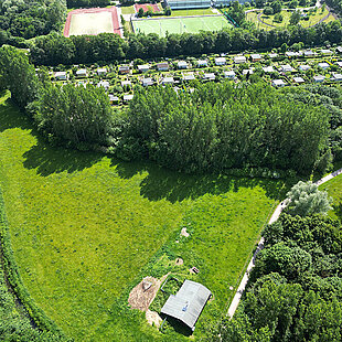 Luftaufnahme eines Parks mit großen Wiesenflächen, einem Gehweg und einem kleinen Gebäude, umgeben von Bäumen und einer Gartenanlage im Hintergrund