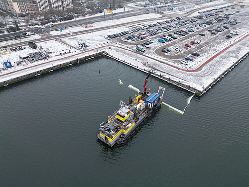 Schiff mit Kran auf Wasserfläche vor schneebedecktem Hafen mit Parkplätzen und Gebäuden im Hintergrund