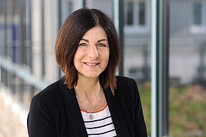 Frau mit schulterlangen, dunklen Haaren und einem lächelnden Gesicht, trägt eine schwarze Jacke und ein gestreiftes weißes Oberteil.