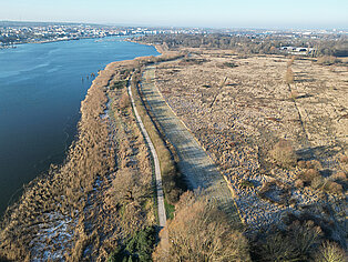 Luftaufnahme der Unterwarnow mit einem Weg entlang des Wassers und einer offenen, teils bewachsenen Fläche rechts